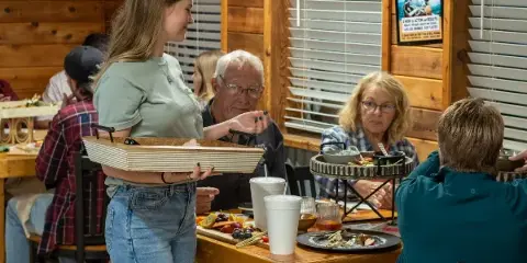 A server carrying a tray in a wood-paneled restaurant dining room with seated guests and food on the table.