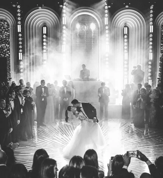 A black and white photograph of a wedding reception featuring a DJ on stage and a couple dancing under dramatic lighting.