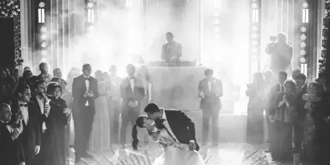 A black and white photograph of a wedding reception featuring a DJ on stage and a couple dancing under dramatic lighting.