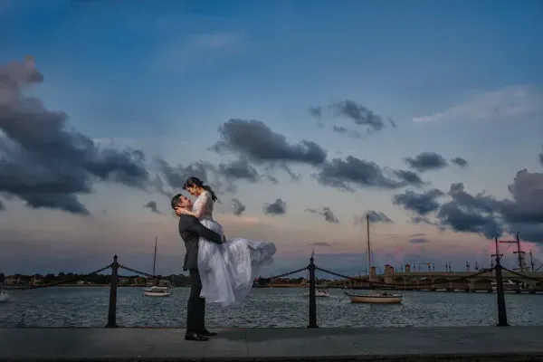 A groom in a dark suit lifts a bride in a white wedding dress at a waterfront promenade with a bridge and boats in the background during twilight.