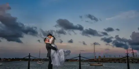 A groom in a dark suit lifts a bride in a white wedding dress at a waterfront promenade with a bridge and boats in the background during twilight.