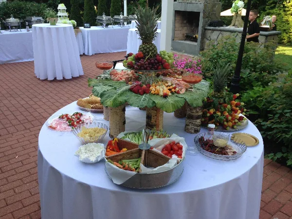 Outdoor catering table with a fruit and shrimp centerpiece, vegetable trays, and a wedding cake in the background.