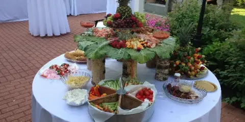 Outdoor catering table with a fruit and shrimp centerpiece, vegetable trays, and a wedding cake in the background.