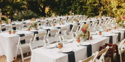 Outdoor event setup with rows of white folding chairs and tables covered in white linens and black runners.