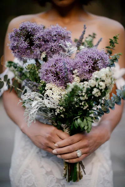 A bride holding a wedding bouquet featuring purple alliums, white flowers, and greenery.