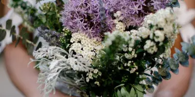 A bride holding a wedding bouquet featuring purple alliums, white flowers, and greenery.