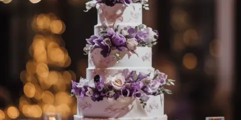 A four-tier white wedding cake decorated with purple and white flowers, displayed on a silver stand with candles in the background.