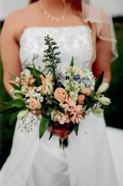 A bride in a white wedding dress holding a hand-tied bouquet of peach roses, white lilies, and blue delphiniums.