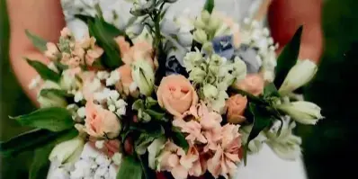 A bride in a white wedding dress holding a hand-tied bouquet of peach roses, white lilies, and blue delphiniums.
