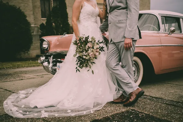 A bride in a white lace gown holding a bouquet and a groom in a grey suit standing next to a vintage pink car.