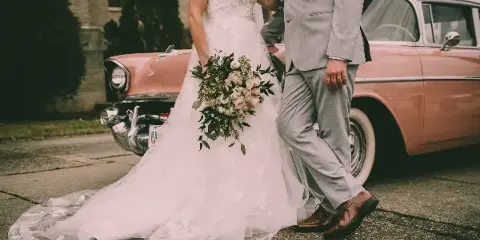 A bride in a white lace gown holding a bouquet and a groom in a grey suit standing next to a vintage pink car.