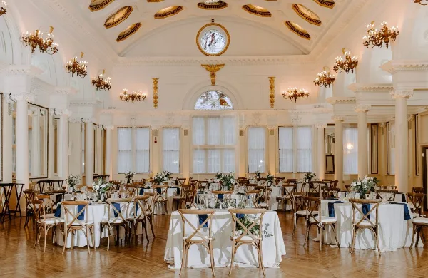 A grand ballroom set up for a wedding reception with round tables, white tablecloths, and wooden chairs.