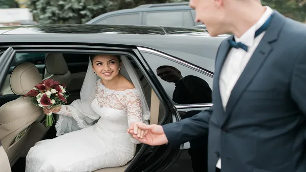 A bride in a white lace gown holding a bouquet sits in the back seat of a black luxury car while a groom in a suit holds her hand.