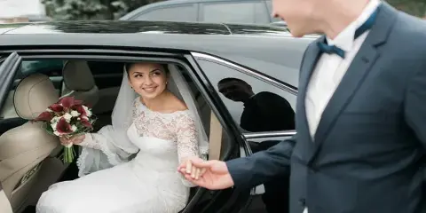 A bride in a white lace gown holding a bouquet sits in the back seat of a black luxury car while a groom in a suit holds her hand.