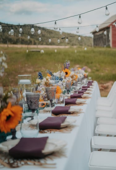 A long event table set with white linens, maroon napkins, floral centerpieces, and string lights in an outdoor setting.