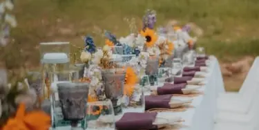 A long event table set with white linens, maroon napkins, floral centerpieces, and string lights in an outdoor setting.
