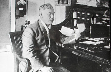 Black-and-white historical photograph of a man in a suit seated at a desk with papers and a filing cabinet in the background.