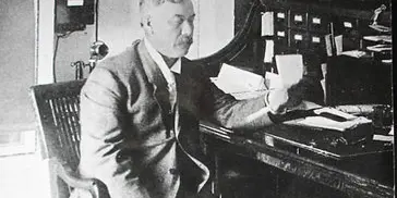 Black-and-white historical photograph of a man in a suit seated at a desk with papers and a filing cabinet in the background.