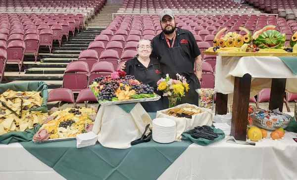 Two catering staff members in black aprons standing behind a table of food platters and snacks in an event hall.