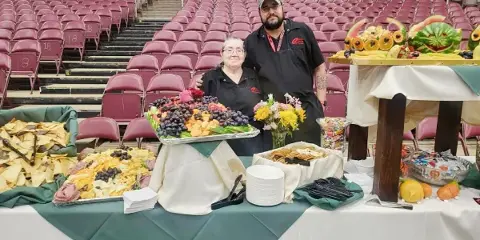 Two catering staff members in black aprons standing behind a table of food platters and snacks in an event hall.