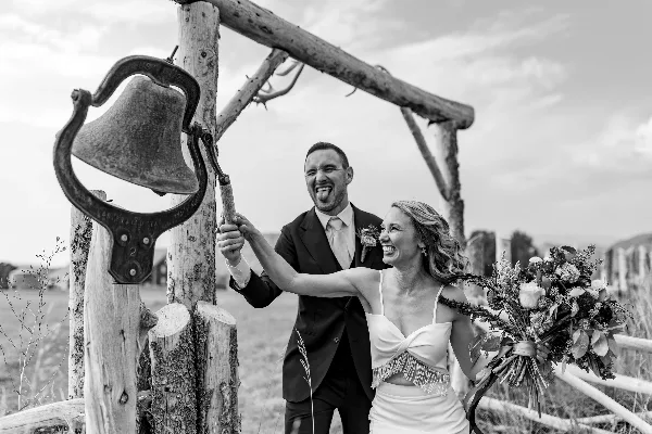 A black and white photograph of a bride and groom laughing while ringing a large bell at a rustic outdoor wedding venue.