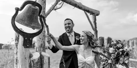 A black and white photograph of a bride and groom laughing while ringing a large bell at a rustic outdoor wedding venue.
