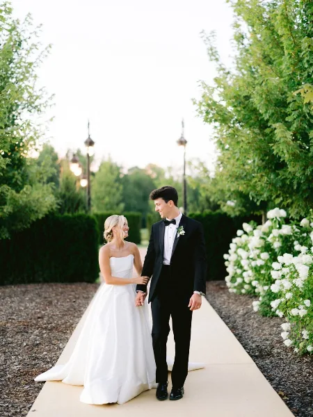 A bride in a white gown and a groom in a tuxedo standing together on a garden path.