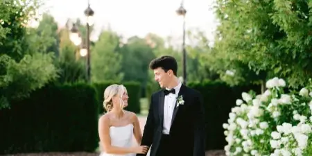 A bride in a white gown and a groom in a tuxedo standing together on a garden path.