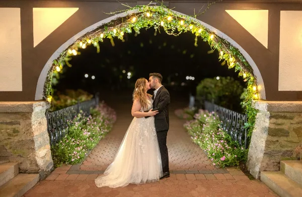 A bride and groom sharing a kiss under a decorative archway with string lights and greenery.