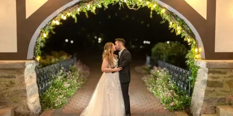A bride and groom sharing a kiss under a decorative archway with string lights and greenery.