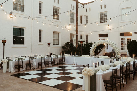 A black and white checkered dance floor and floral arch set up in a white-walled courtyard with string lights and tables.