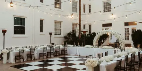 A black and white checkered dance floor and floral arch set up in a white-walled courtyard with string lights and tables.