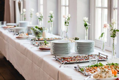 A long table set with a white tablecloth, featuring stacks of white plates, floral centerpieces, and various food dishes on silver trays.