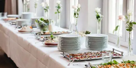 A long table set with a white tablecloth, featuring stacks of white plates, floral centerpieces, and various food dishes on silver trays.