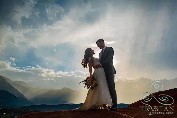 A bride and groom standing on a rocky outcrop with a mountain landscape and dramatic sky in the background.