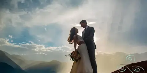A bride and groom standing on a rocky outcrop with a mountain landscape and dramatic sky in the background.