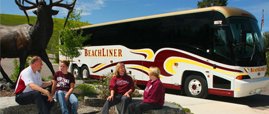 A large white and gold BeachLiner charter bus parked outdoors with a group of people sitting in front of it.