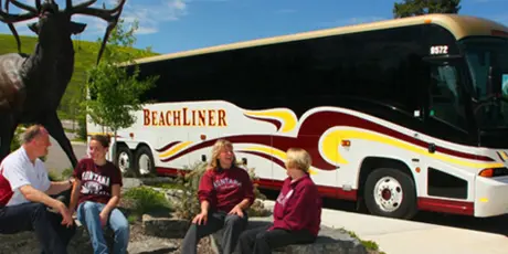 A large white and gold BeachLiner charter bus parked outdoors with a group of people sitting in front of it.