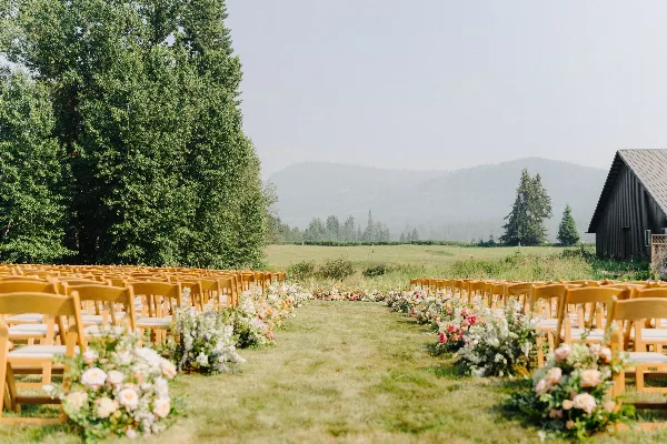 Outdoor wedding ceremony setup with rows of wooden chairs and floral arrangements in a grassy field with mountains in the background.