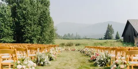 Outdoor wedding ceremony setup with rows of wooden chairs and floral arrangements in a grassy field with mountains in the background.
