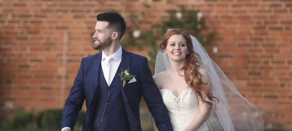 Bride and groom in wedding attire standing together in front of a brick wall