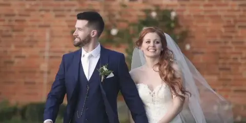 Bride and groom in wedding attire standing together in front of a brick wall