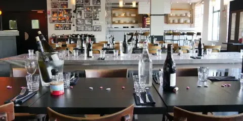 Interior view of a restaurant dining room with wooden tables, chairs, and a bar area in the background.