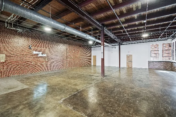 A spacious, empty event hall with exposed brick walls, polished concrete floors, and a neon sign that reads 'The Venue Cotton Creek'.