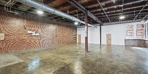 A spacious, empty event hall with exposed brick walls, polished concrete floors, and a neon sign that reads 'The Venue Cotton Creek'.