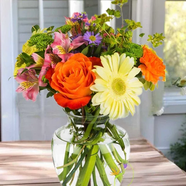 A colorful mixed bouquet featuring orange roses, yellow gerbera daisies, and pink alstroemeria in a clear glass vase.