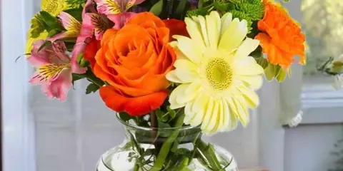 A colorful mixed bouquet featuring orange roses, yellow gerbera daisies, and pink alstroemeria in a clear glass vase.