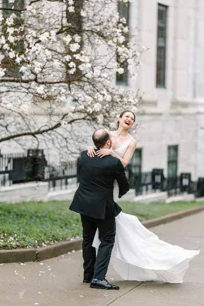 A bride and groom laughing together outdoors near a blooming tree.
