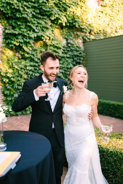 A bride and groom laughing and holding champagne flutes at an outdoor wedding reception.