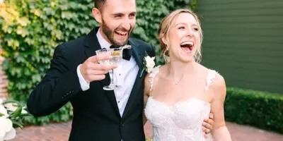 A bride and groom laughing and holding champagne flutes at an outdoor wedding reception.